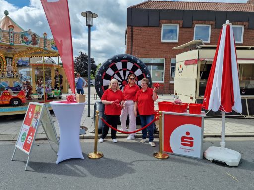 Drei Personen in roten T-Shirts stehen vor einem Dartscheiben-Stand bei einer Veranstaltung.