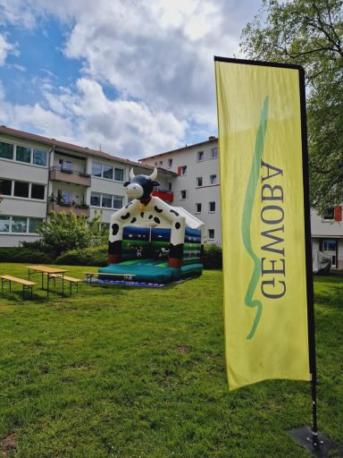 Gelbe Fahne mit Aufschrift "CEMOBY" vor einem bunte Hüpfburg und Wohngebäuden.