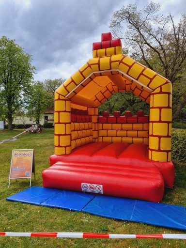 Bunte Hüpfburg in Form einer Burg auf einer Wiese, umgeben von Bäumen.