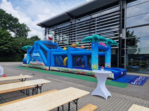 Bunte Hüpfburg im Freien mit Palmen, Tischen und Stühlen im Hintergrund.