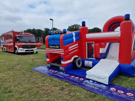 Luftige Hüpfburg in Form eines Feuerwehrautos auf einer Wiese, neben einem Einsatzfahrzeug.