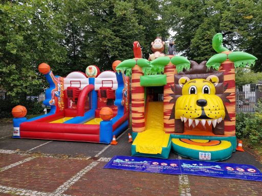 Buntes Hüpfburg-Set mit Basketballkorb und Löwenrutsche, umgeben von Bäumen.